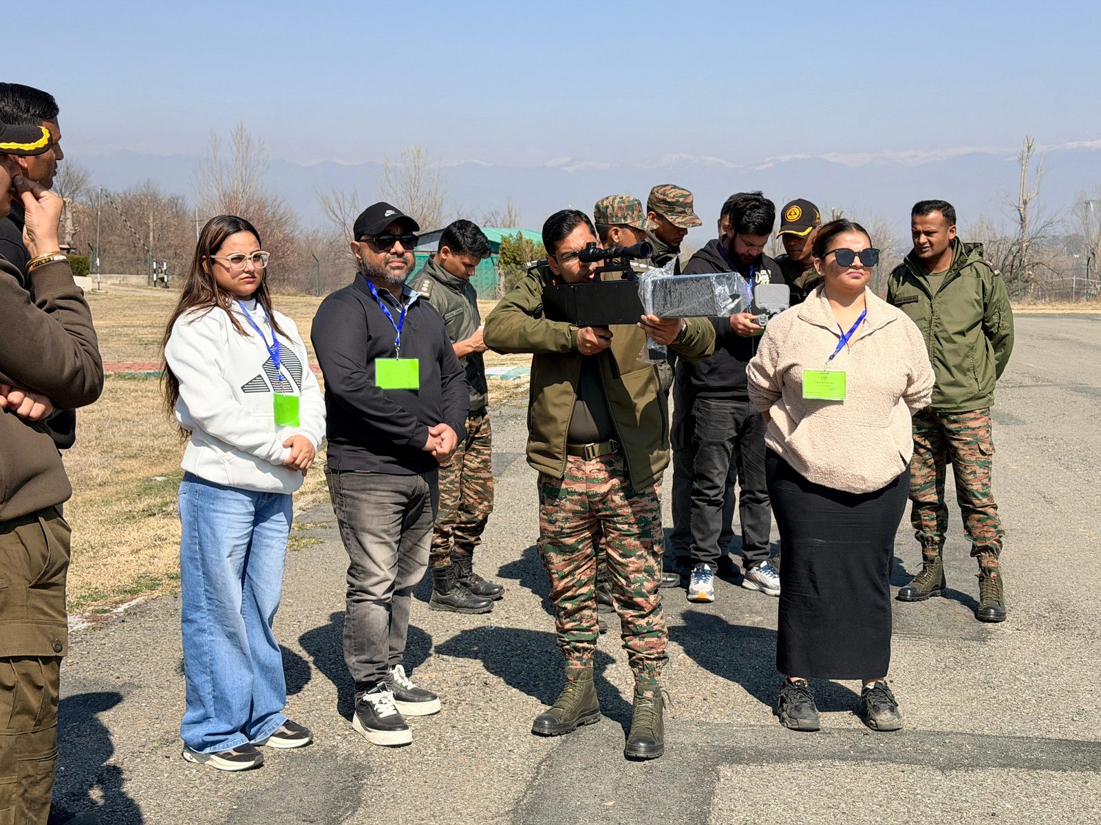 Officers and civilians observing a soldier aiming a handheld anti-drone jammer in mountain terrain